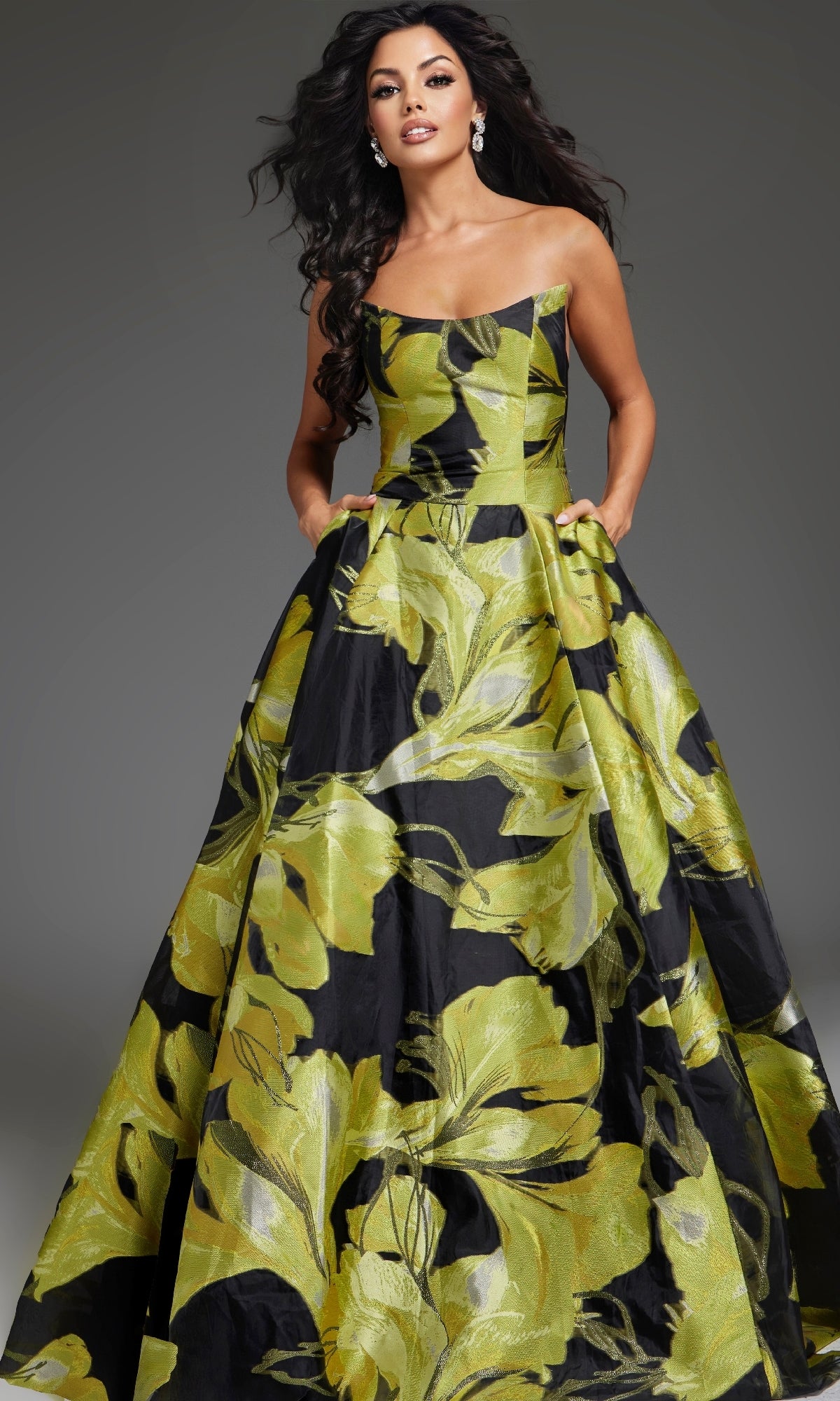 A woman poses with hands in pockets wearing the Jovani 42435, a strapless long prom dress featuring bold green floral patterns, set against a neutral background.